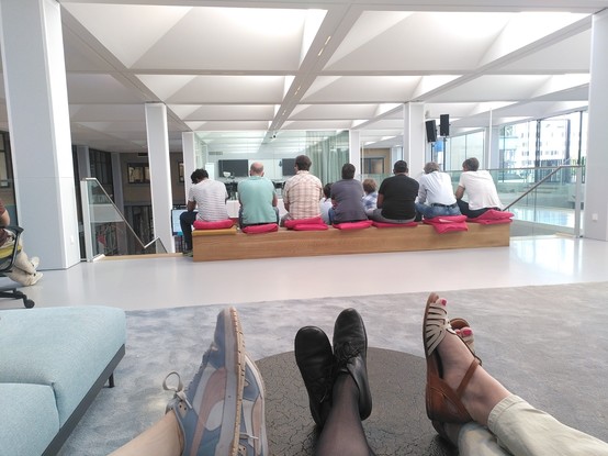 Three pairs of feet on a table. Further away 7 people are sitting on a bench to watch a presentation.