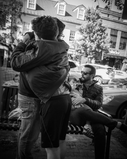 Black and white photo of two people hugging on a sidewalk. Their faces are not visible. One of them is hugging the other so hard, the seem to be slightly lifting them off the ground a little: their jacket has been scrunched up a bit. Just behind and to the right of this duo, a bearded man wearing sunglasses cradles a boy in his arms as they sit on a bench. Beyond in the background are passing cars, trees, building on the other side of the street.