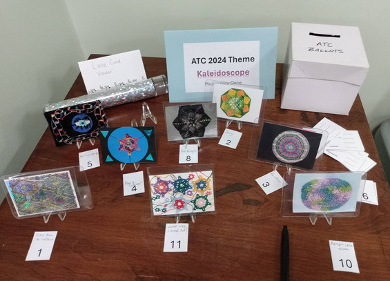 A table that shows our ATC 2024 Theme as Kaleidoscope. There are 8 entries this year. Some are bobbin lace, some are needle lace, some tatting, or crochet lace.

They have repetitive motifs typically, some with eyeball images in the center. Some have shiny foil backgrounds. Each has an artwork title and a number.

There's a ballot box in the back and the ballot forms with the category list. 

Possible category votes are: most unusual'; most technically impressive; best representation of the theme; and favorite ATC.