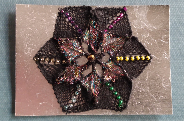 A six-pointed star-like object with  black linen diamonds around the outer edge. In the center of each diamond shape motif is a single line of colored glass beads. One is pink, one is purple, one is like a gold, one is aqua, one is green, one is yellow.

In the center of the star is a set of 6 smaller diamonds that are made with metallic thread so they are all shimmery and multi-colored. They are sort of wrapped with thread on the lines closer to the center. 

In the very center is a small single gold bead that is kind of like a disco ball. 

ATC cards are 2.5 x 3.5 inches so you have to fit into that space.