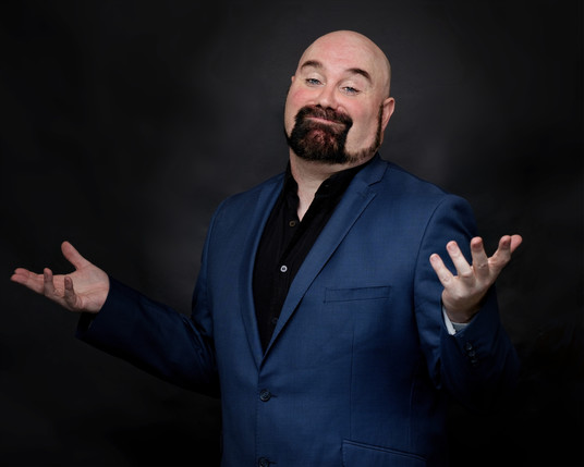 Actor Matt Whitfield in a blue suit with black shirt with his arms outstretched, palms up. He has a shaven head, black beard and beautiful blue eyes.