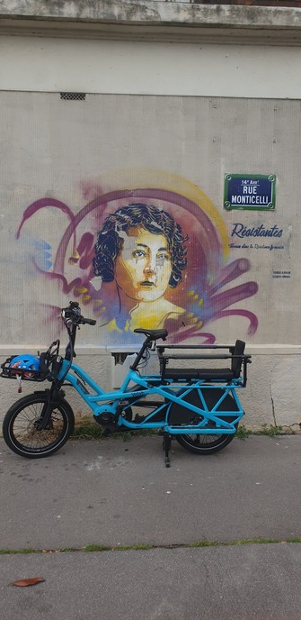 Une fresque sur un mur représentant une femme. A côté une inscription Resistantes et la plaque de la rue. Devant le mur un vélo bleu