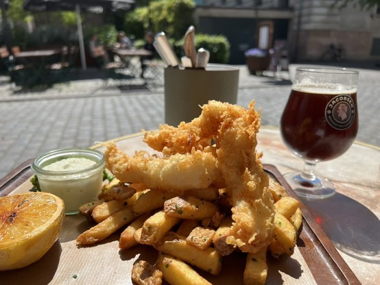 A plate of crispy fried fish skewered on a stick and seasoned french fries, accompanied by a creamy dipping sauce and a grilled half lemon, served on a wooden board with a glass of dark amber beer in the background, all set on an outdoor table in a sunny cafe setting with greenery and patrons enjoying their meals.