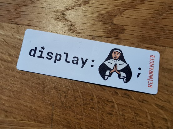 White sticker on a wooden table with a CSS declaration on it stating the display property with an emoji of a nun as value