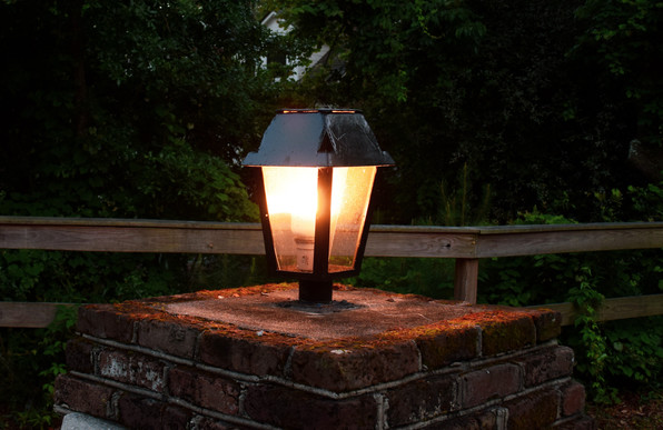 A close up photo of a lamp atop a brick pedestal in late evening.  The lamp has a rectangular black frame and top.  