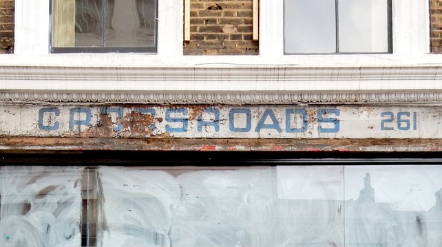 Shopfront with a fading and damaged sign that has 'Crossroads' in light blue on white in digital-style letters with curved terminals. To the right of this is the building number, 261, painted in the same blue.