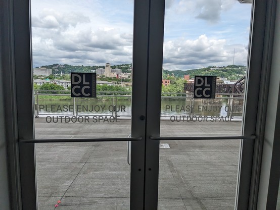 Doors that read "please enjoy our outdoor space" and have a beautiful view of water, bridges, and distant buildings, greenery, and clouds