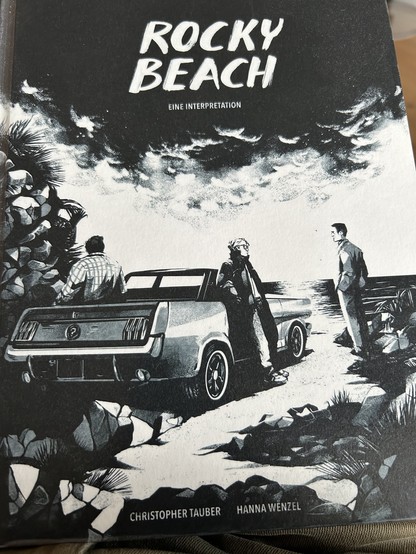 Cover der Graphic Novel „Rocky Beach“. Schwarzweiß Zeichnung: Drei Männer stehen oder lehnen an einem Auto, das am Meer parkt