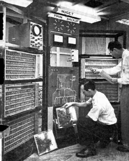 old computer in a server room being worked on by two individuals in black and white.