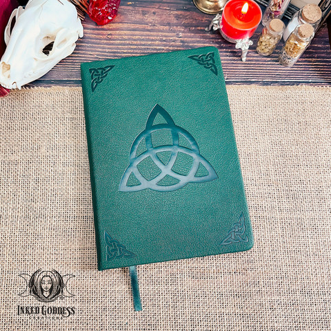 An emerald-green journal is lying on a burlap-covered surface. The cover has a leather texture with a large Celtic triquetra, also known as a tri-knot, debossed in the center. Each corner also has a debossed Celtic knot, and a green ribbon is attached to the spine to be used as a bookmark. This is an exclusive product from Inked Goddess Creations.