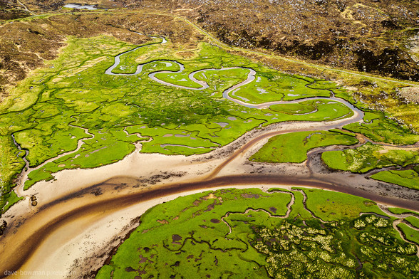 Experience the vast expanse of Harris Marshland, situated on the Isle of Harris in the Outer Hebrides, Scotland. A mosaic of vibrant greens stretches into the distance, interrupted only by winding water channels meandering towards the sea. This aerial perspective captures the intricate beauty of nature's design, where land and water converge harmoniously in a serene landscape.