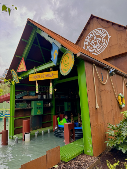 A river rapids boat enters a themed mine shaft featuring bright colours and warning signs. Overhead, the “Amazonia Mining Company” logo is seen on a building. 