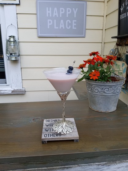 A milky light violet substance in a martini glass with a pick stabbing three blueberries. The glass stem is a series of vines and leaves in steel. The glass is on a wood coaster shaped like a pallet. The coaster reads, 