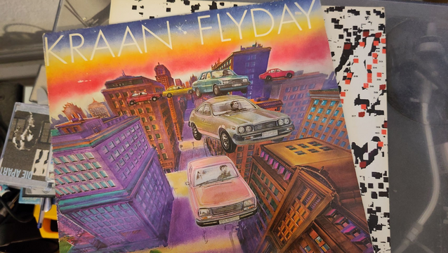 Top down photography. It shows the front cover of Kraan's album "Flyday". Very colourful. It displays some 70s and 80s car models flying through a neighbourhood of tall 10 stories high stone buildings. Please ignore some of the cables and unsorted tapes caught on camera at the borders of the photo. 