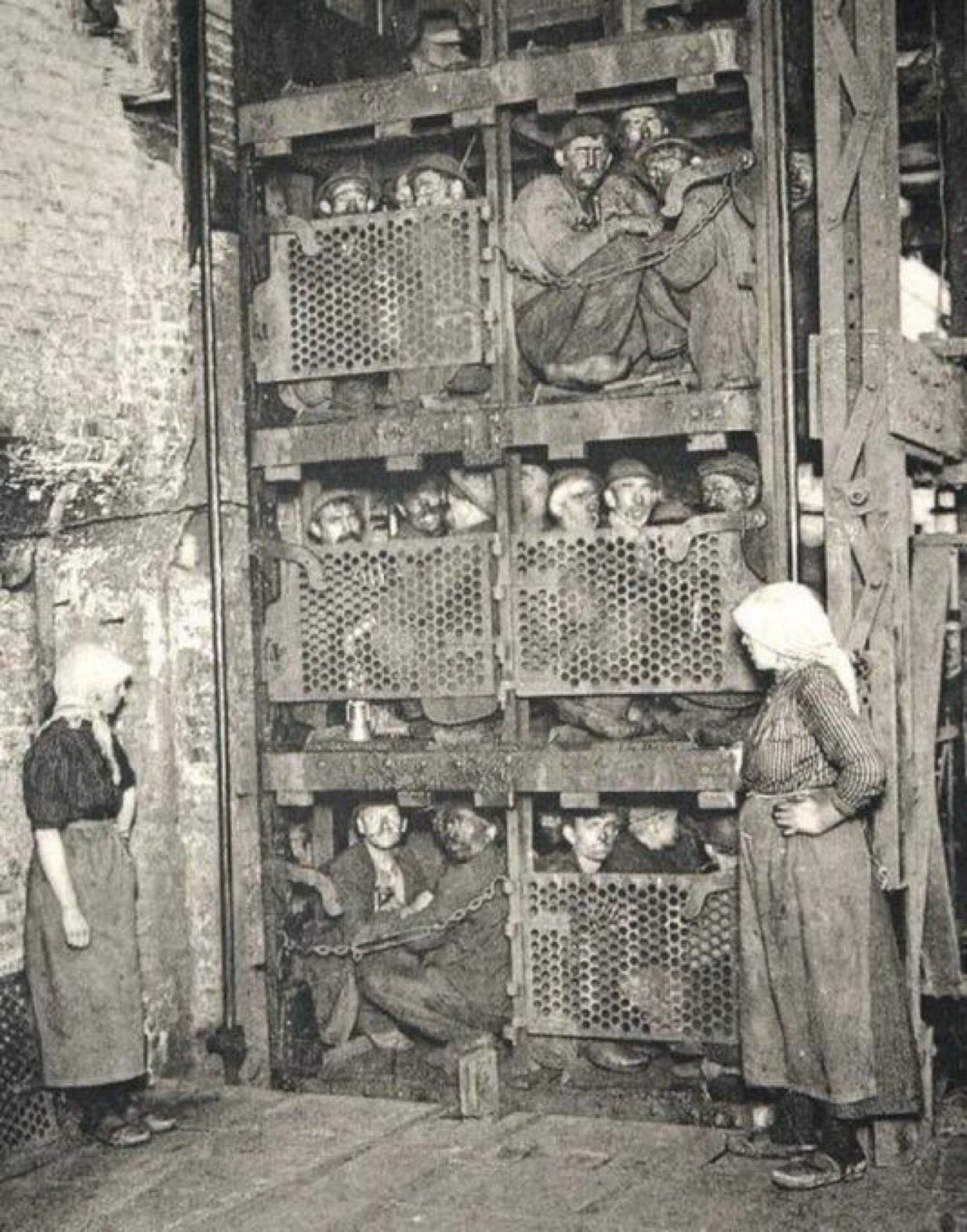 Une photographie historique en noir et blanc montre des mineurs entassés dans une cage d'ascenseur à face métallique. L'ascenseur est dans un cadre industriel, et deux femmes, debout à proximité, observent la scène.