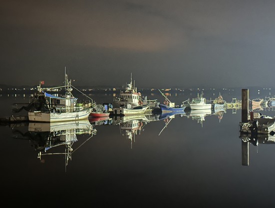 The photo shows a group of fishing boats docked in a harbour at night, in Costa Nova. The boats are all different shapes and sizes and are tied up to the dock with lines. Some boats have lights on, casting a warm glow on the water. In the background, you can see the outline of buildings against the night sky.