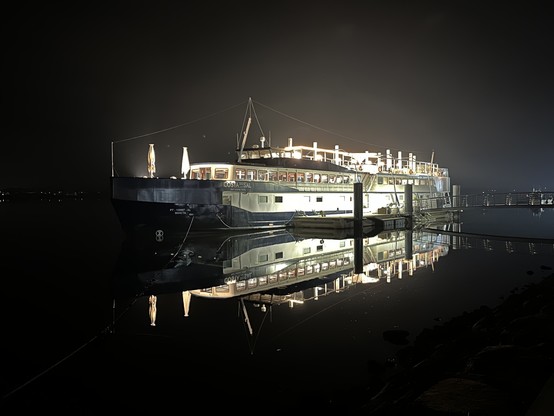 The image features the Costa do Sal, a docked ship transformed into a hotel and restaurant in Costa Nova, Portugal. The name Costa do Sal translates to 'Coast of Salt.' The boat is illuminated at night, with warm yellow lights casting a welcoming glow on the water's surface.  You can likely make out the outline of decks and cabins on the ship. In the background, the darkness reveals the faint shapes of buildings lining the shore.
