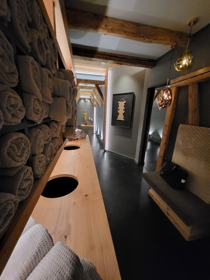 A room with a bench and towels is depicted in this image. The room has a black color scheme with brown accents, featuring a bench against the wall. There are towels neatly rolled up on the bench, adding a touch of luxury to the space. The room has a vase and a lampshade, suggesting a cozy and well-designed interior. The flooring appears to be hardwood, and there is a mirror on the wall. The overall atmosphere of the room is elegant, with a hint of a hotel-like ambiance.