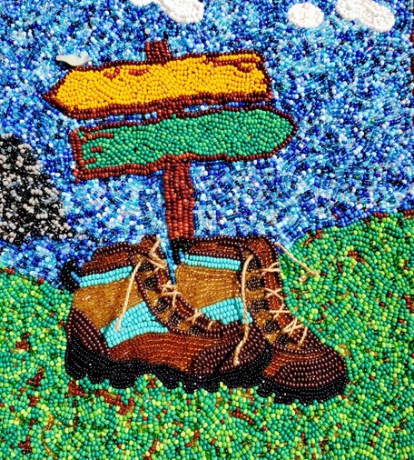 Bead emboidery. A pair of brown and turquoise boots sitting un the grass under a green and yellowish trail sign. Blue sky with a few clouds  above. Gray mountain far back on the left.