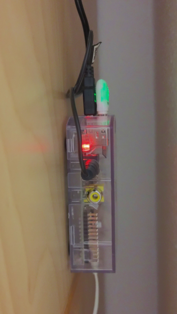 Foto eines sich im Betrieb befindlichen Raspberry Pi Model B in einem transparenten Gehäuse. Zusätzlich zum MicroUSB-Stecker für die Stromversorgung stecken ein WiFi-USB-Adapter und ein USB- und Klinkenstecker von Lautsprecherboxen (nicht sichtbar) in dem Gerät. Das Raspberry Pi-Gehäuse klebt am Seitenteil eines Schranks und steckt zwischen diesem Schrank und einer Wand in einer Nische.