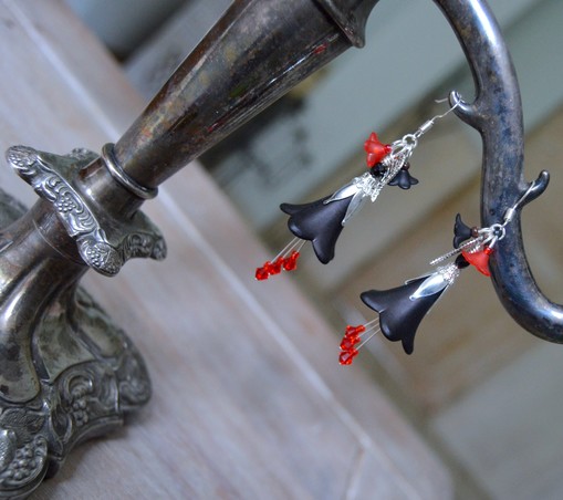 A pair of black flower shaped earrings with scarlet red crystals hanging underneath. They hangs from a vintage silver Victorian candelabra on a white table.