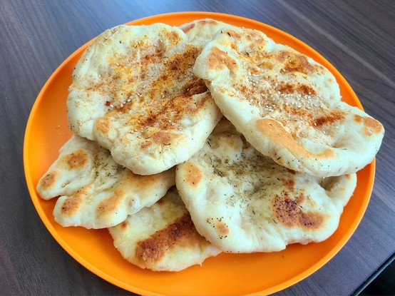 Several pieces of lumpy (in a good way) flatbread piled up on an orange plate. Some of the flatbreads have paprika and sesame seeds, while the rest have garlic and Italian seasoning. The bread is whitish with golden-brown spots from where it was touching the pan.