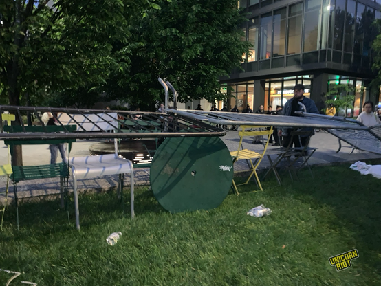 Metal barricades laid over overturned lawn chairs sideways not as intended by police presumably as part of protest barricade. Evening dusk lighting over concrete and dark green grass. Trees and campus building seen in background round