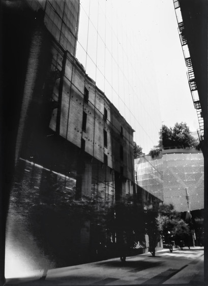 A black and white pinhole photo of a glass office building reflecting an old brick warehouse