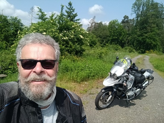 Ein älterer Mann mit Sonnenbrille, dahinter ein Motorrad. Im Hintergrund Bäume und Sträucher.