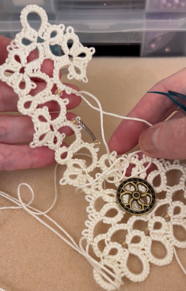 A piece of unfinished handmade lace in tatting. Thick thread in cream white color with a metal button resembling a mechanical wheel. Paper clips are temporarily holding beads in place until the lacework reaches them.