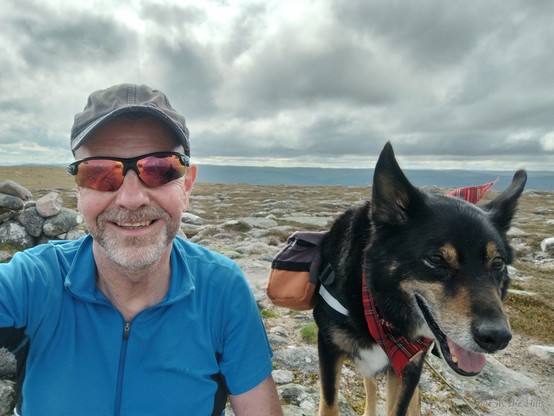 Selfie of me and Moray the dog, sat next to a cairn on a hillside with some rocks.