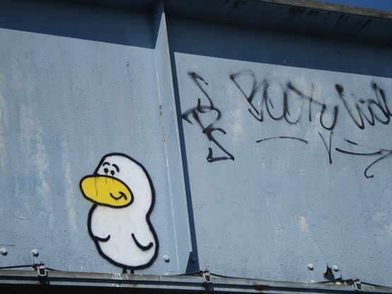 A spray painted pseudo duck-like character on the side of a grey painted steel railway bridge panel. White with yellow beak and small stumpy wings with a mouth that suggests innocent contentment. Black written graffiti containing the word Booty is beside it.