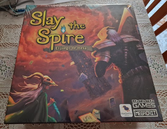 Caja del juego de mesa de Slay the Spire. Donde se puede ver a una asesina armada con un cuchillo y un guerrero equipado con una espada, ambos de espaldas. Al fondo se puede ver la torre/espiral rodeada de nubes naranjas.