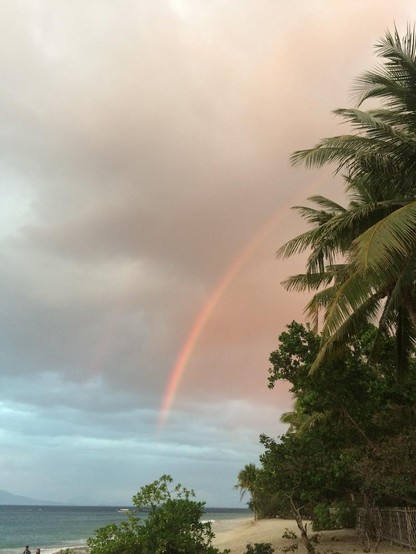 Philippines, beach with lots of palmtrees, dark clouds with a partly rainbow coming from behind the palmtrees, shooting down towards the sea. 