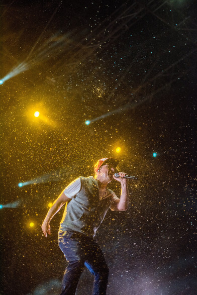 The image captures a dynamic moment during a live musical performance. The scene is filled with energy and excitement, as the performer, a man wearing a cap, is caught in the midst of singing into a microphone. He is dressed in a casual outfit consisting of a vest over a t-shirt and jeans.

The stage lights create a dramatic effect, with bright yellow and blue beams cutting through the darkness. These lights illuminate particles in the air, possibly mist or smoke, adding a textured and vibrant backdrop to the performer. The overall mood of the image is intense and lively, conveying the high-energy atmosphere of a live concert. The performer appears deeply immersed in the music, contributing to the captivating and electrifying ambiance of the scene.