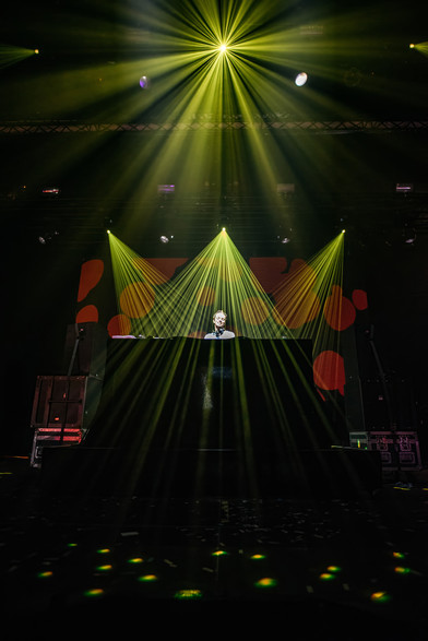 The image captures a DJ performing on stage, enveloped in a dramatic and vibrant light show. The DJ stands behind a large console, wearing headphones, focused on their music. The stage is illuminated by numerous beams of yellow and green light radiating from above and behind the DJ, creating a visually stunning display that adds energy and excitement to the scene.

The background features a screen with abstract patterns and designs, further enhancing the dynamic atmosphere of the performance. The lights cast intricate patterns on the stage floor, adding to the immersive experience. The overall mood is electrifying and energetic, typical of a lively DJ set at a concert or music festival. The combination of the DJ's concentration and the spectacular lighting creates a powerful visual and auditory experience for the audience.