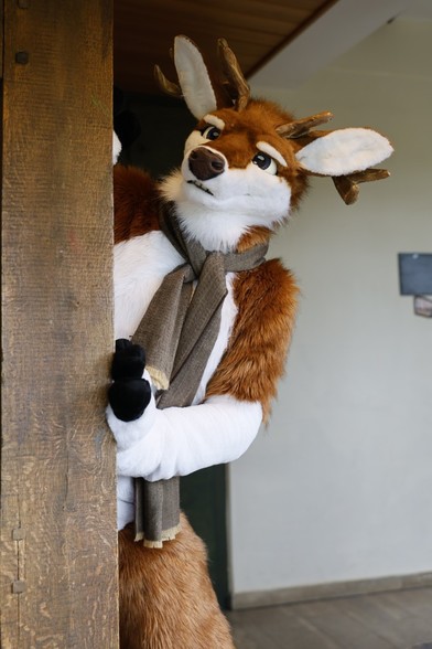 A red deer fursuiter wearing a scarf peeping from behind a wooden pillar