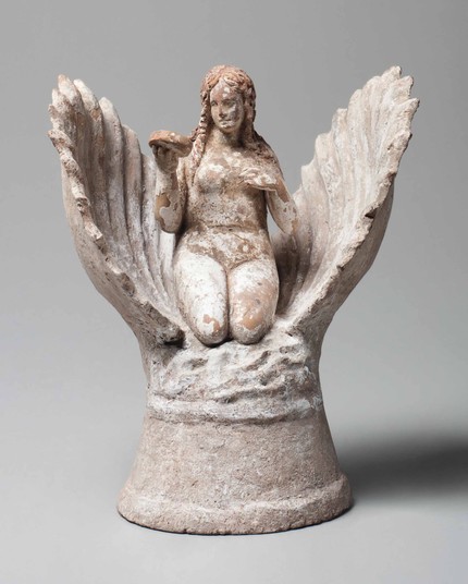 Photo of a terracotta figurine depicting the birth of Aphrodite. The goddess emerges from an open scallop shell, depicted nude, seated with her legs tucked beneath, her long wavy hair falling onto her shoulders, looking at her reflection in a mirror held in her right hand, on a high cylindrical plinth. Some polychromy and white slip are preserved throughout. The nude goddess is raising her right hand holding her mantle, a dolphin below to her right, her lowered left hand on a column, preserving…