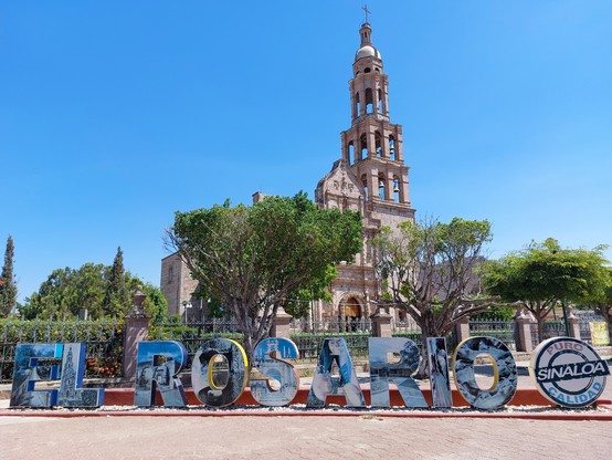 Blue sky, ancient Spanish era church with an almost open bell tower, green trees and colorful letters in the front with the name of the city "El Rosario" in Sinaloa, Mexico. 
