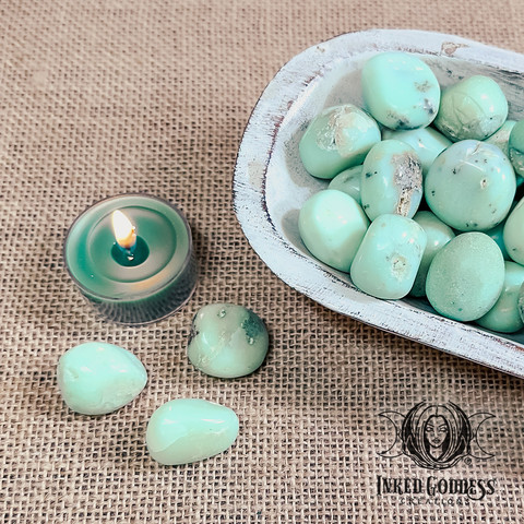 Three specimens of the "Mint Chrysoprase Tumbled Gemstone" product are arranged on the left in front of a tealight candle. Several more specimens fill a whitewashed wood bowl on the right. Presented by Inked Goddess Creations.
