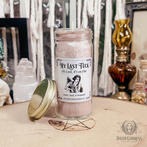 A mini jar candle rests on a wood slice. It's thinner than a basic jar candle. The jar is made of glass and the candle itself is a frosty pink color. The brass screw-on lid is resting against the base of the jar. The label on the glass reads, "My Last Fuck," "Oh Look, It's on Fire". This is an exclusive product from Inked Goddess Creations.