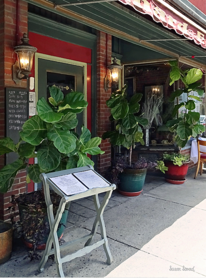 One of Kingston NY's quaint restaurants with outdoor seating decorated with green foliage in decorative pots. A folded white stand holding a menu is placed in front, inviting passersby to explore the culinary offerings.