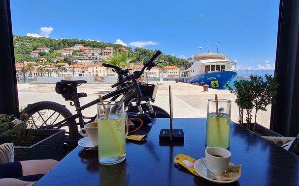 Coffee table with two cups of coffee and two glasses of vitamin drink. In the background are bicycles and a ship in the sea and a hill with houses.