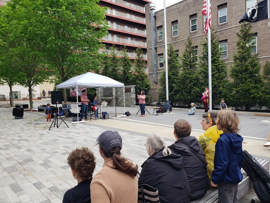 A student performs outside with a red electric guitar, standing underneath a canopy. Closer to the camera are onlookers, sitting in a row.