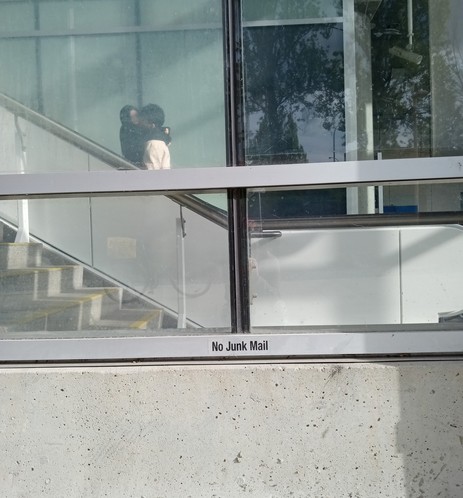 couple kissing on escalator through window, inexplicable 'no junk mail' label on outside of building