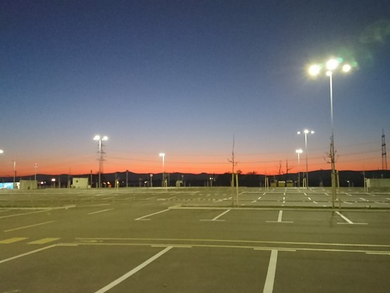 Motiv:
Ein leerer Parkplatz unter einer untergehenden Sonne. Im Hintergrund sind Hügel und eine Stromleitung zu sehen.

Bildformat:
Querformat.

Genre:
Landschaftsfotografie.

Hintergrund:
Im Hintergrund sind Hügel zu erkennen, über denen sich eine Stromleitung erstreckt. Einige Gebäude sind ebenfalls sichtbar.

Farben:
Der Himmel zeigt ein Farbspiel von tiefem Blau bis zu einem leuchtenden Orange am Horizont, während die Lichter des Parkplatzes in hellem Weiß leuchten.

Metapher:
Der leere Parkplatz ruht wie eine Bühne nach dem letzten Akt, während die Sonne sich wie ein stiller Applaus über den Horizont legt.