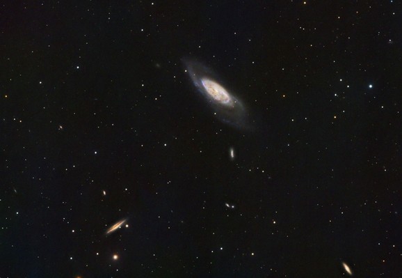 Messier 106:
Black background with stars of varying brightness and colour spread throughout. Star colours are primarily white and shades of orange or blue.

At upper center is a spiral galaxy with a large yellow-shade disk, seen partially edge on. There are fainter outer arms. Some blue and  red patches can be seen in the disk.

There are also some small disk-shaped galaxies visible in the lowe half of the image. There is one lower left that is yellow with a dark lane running through it length-wise. 