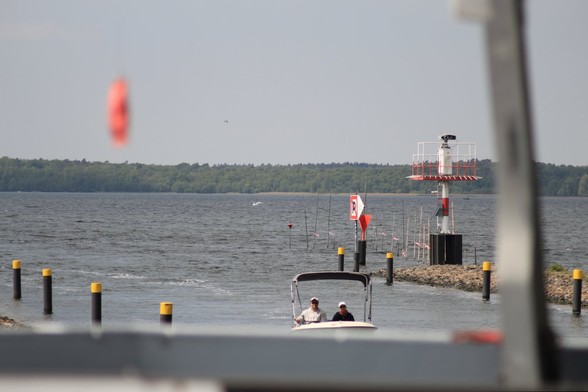 Im Vordergrund die Brüstung eines Schiffs, ander Flußmündung dahinter ein kleines Boot mit zwei Personen. Links und rechts Leitpfosten am Ufer. Dahinter ein See mit Wald am Ufer auf der anderen Seite. Seevögel in der Luft.