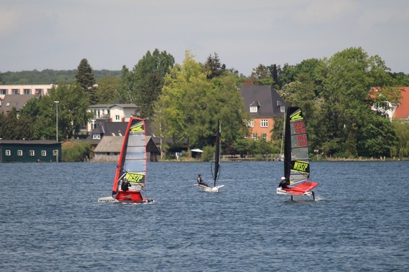 Drei Surfer/ Segler mit Segel in rot, schwarz und gelb. Einer links und
rechts von der Seite, einer in der Mitte frontal.