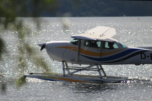 Ein Wasserfulgzeug mit Schwimmern an der Unterseite sitzt auf dem Wasser auf. Farbe weiß, Wellen-Details in gelb, blau und schwarz. In der Kabine sind grob ein paar Leute zu erkennen.